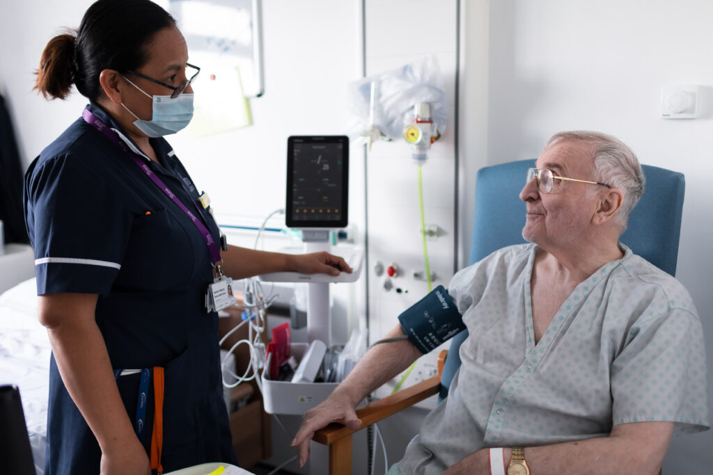 Nurse talking to a male in his late 60s. The man is having his blood pressure checked in hospital.