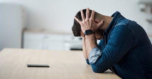 Man looking stressed with head in his hands with his phone on the table near him.