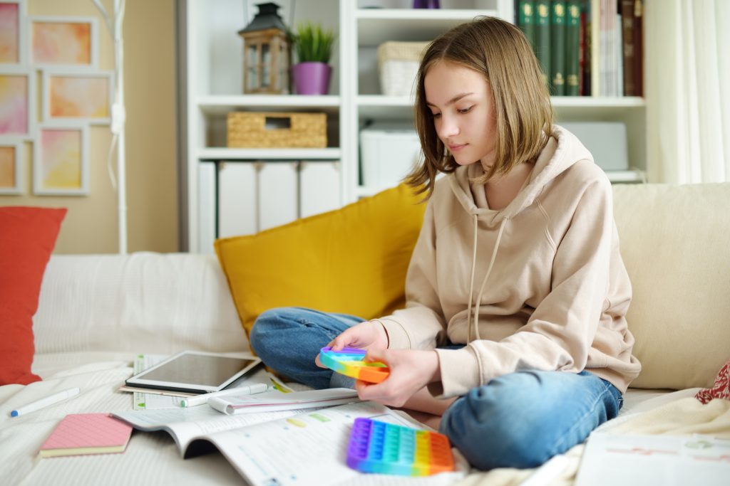 eenage girl playing with rainbow pop-it fidget toy while studying at home. Teen kid with trendy stress and anxiety relief fidgeting game.