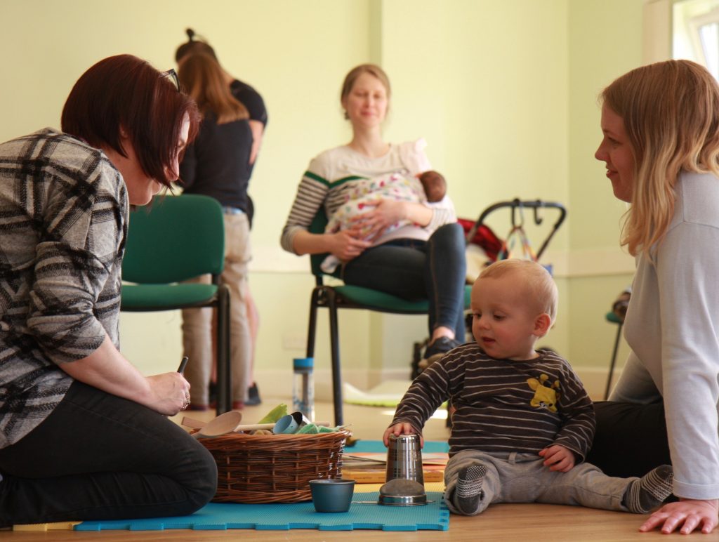 A baby hub with a woman and her baby talking to a health visitor, and a mum breastfeeding in the background.